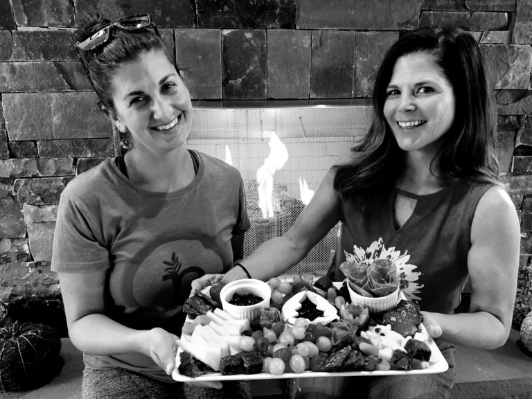 Two women holding a cheeseboard in front of a fireplace.