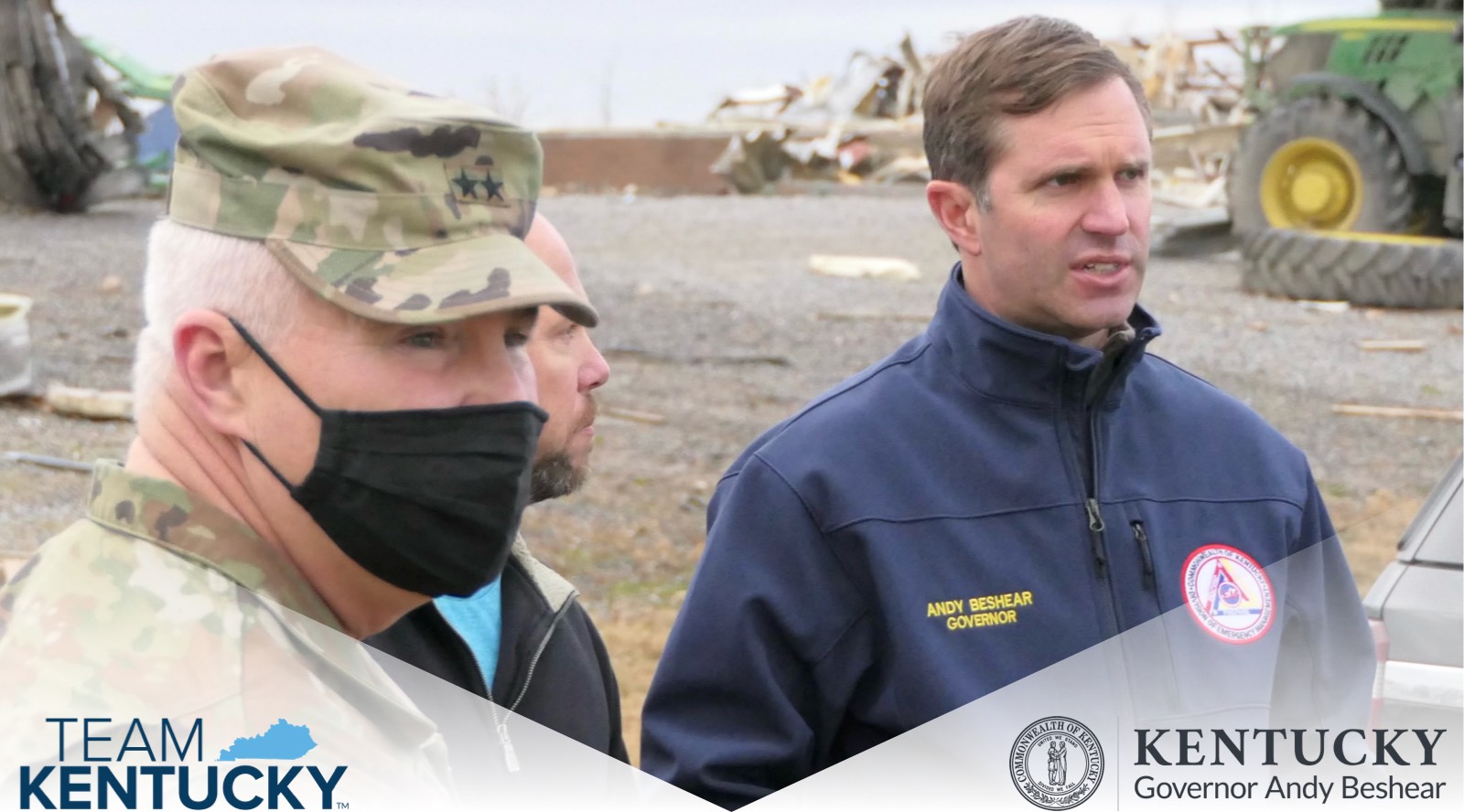 Governor of Kentucky, Andy Beshear, stands with two men, one wearing a military uniform, with tractors in the background.